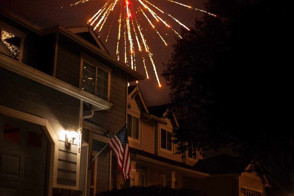 Fireworks exploding over a suburban home with an American flag at night, illustrating holidays when do people pop fireworks